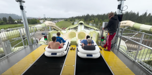 Two groups of people prepare to ride a water slide, with a staff member overseeing from the platform above.